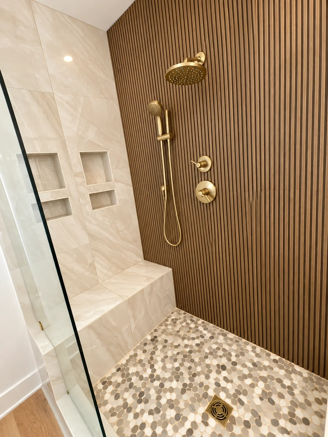 Fox Hill primary shower with a wood slat accent wall, pebble floor, and brass fixtures