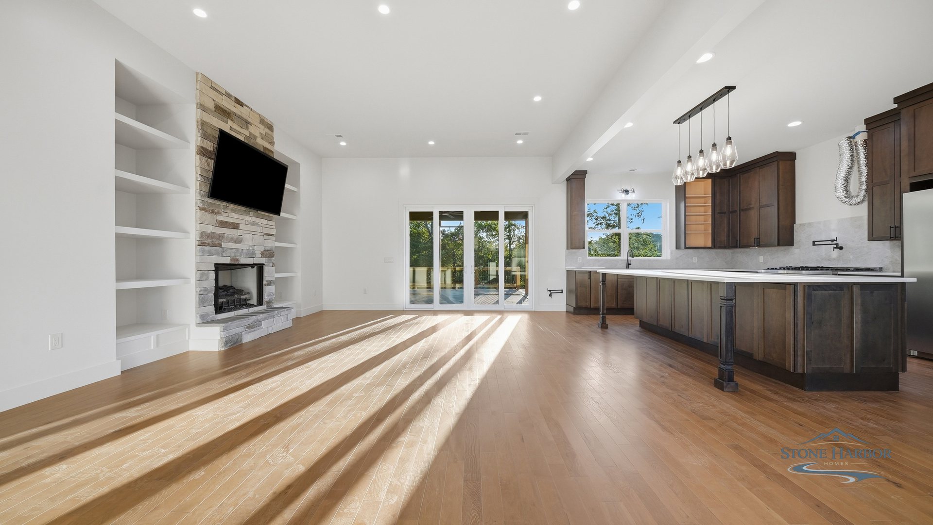 Open great room with a stone fireplace, built-in shelving, and wood floors opening to the kitchen